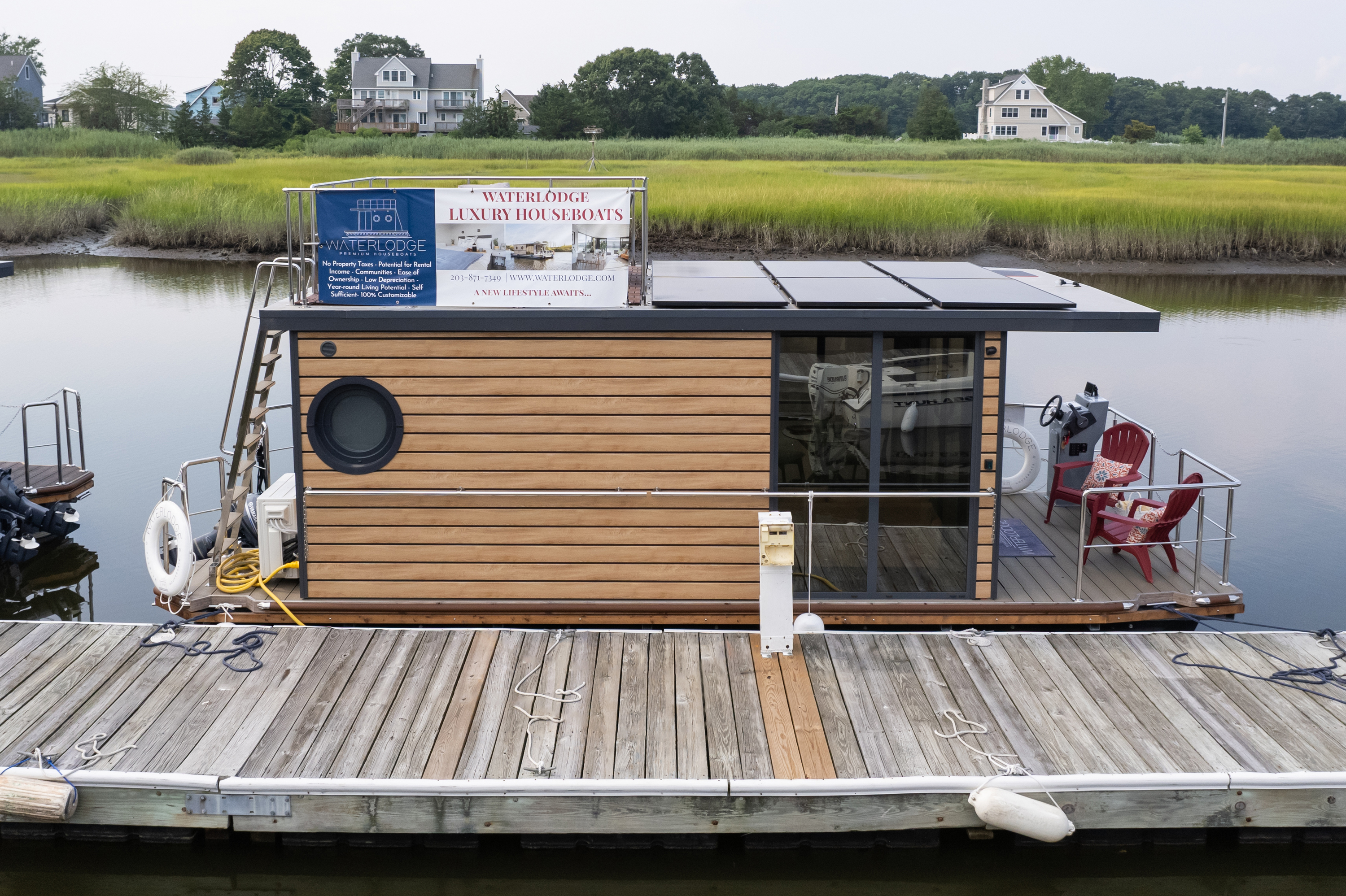 A floating wooden structure with a sign “Waterhole Luxury Houseboats” docked at a pier, surrounded by calm water and greenery.