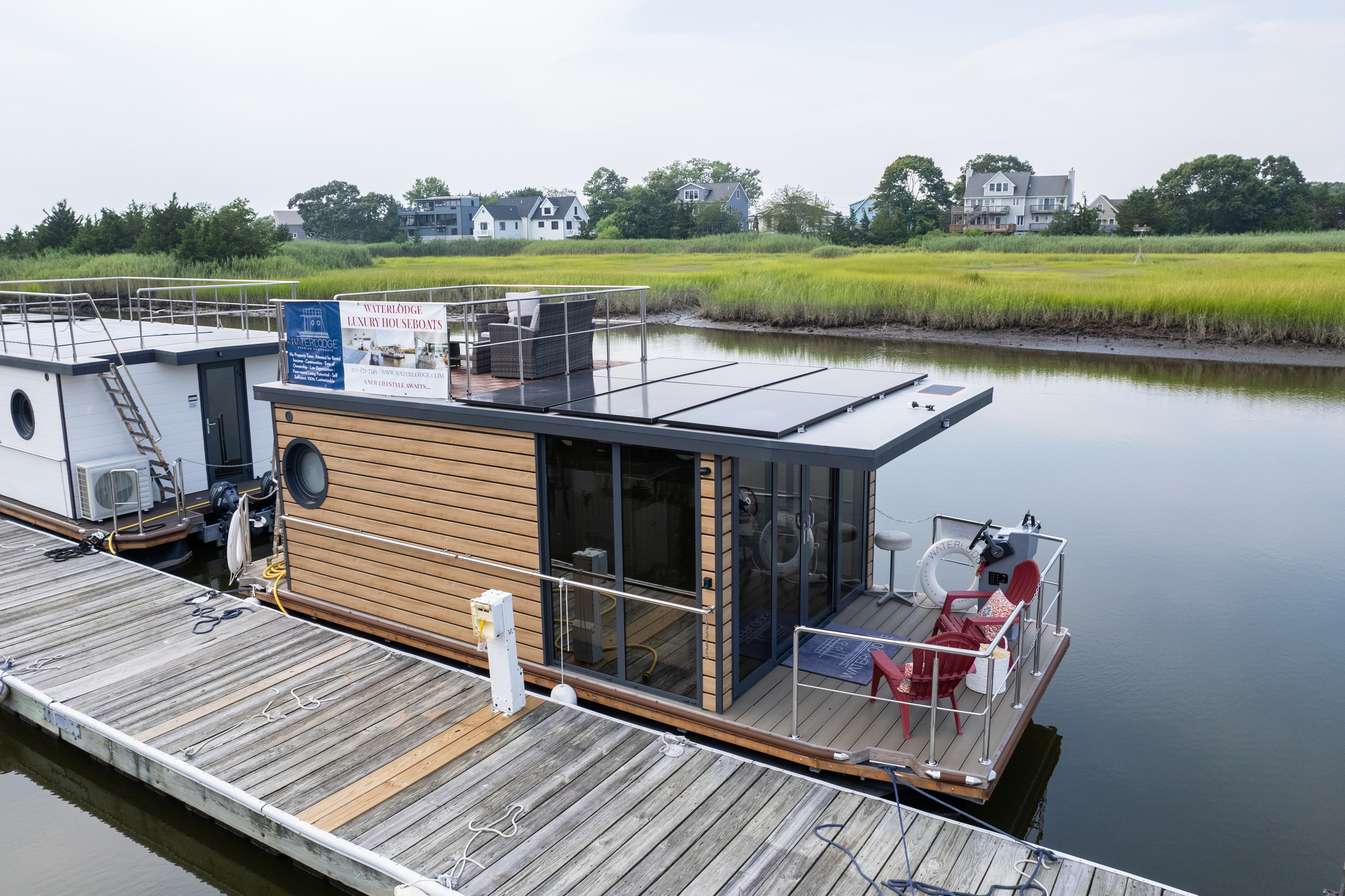 A floating wooden structure with a sign “Waterhole Luxury Houseboats” docked at a pier, surrounded by calm water and greenery