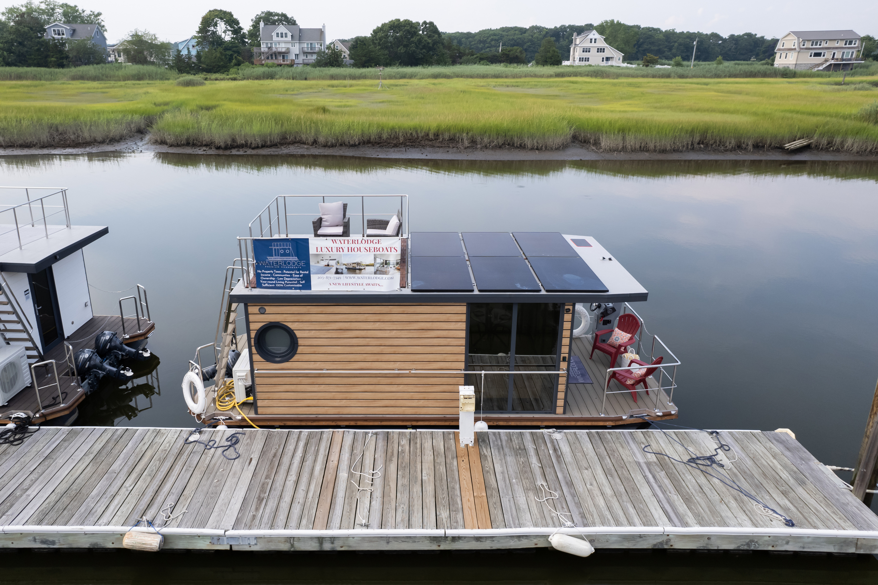 A floating wooden structure with a sign “Waterhole Luxury Houseboats” docked at a pier, surrounded by calm water and greenery.