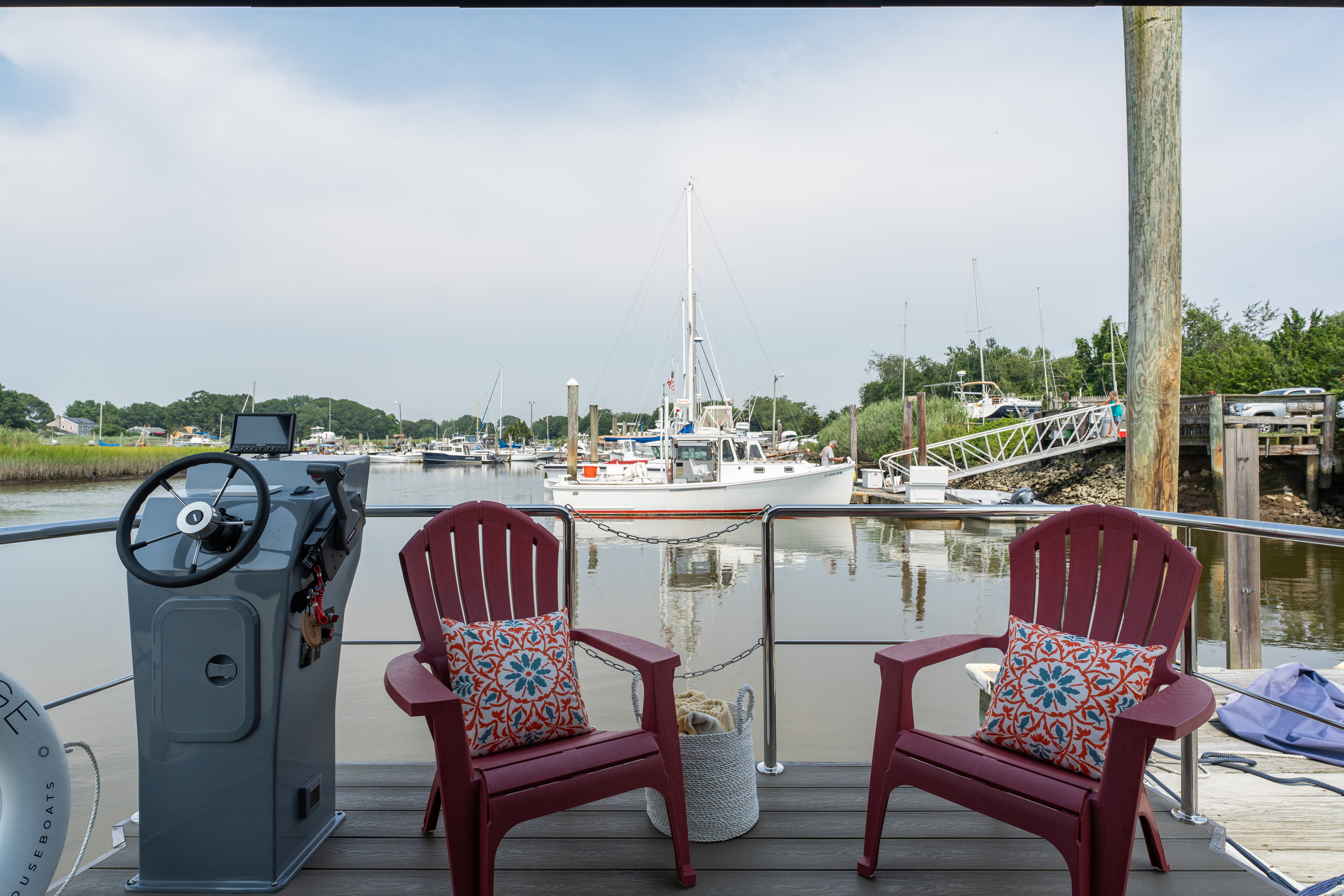 Two red chairs on a dock overlooking a serene marina with boats and lush greenery under a cloudy sky.