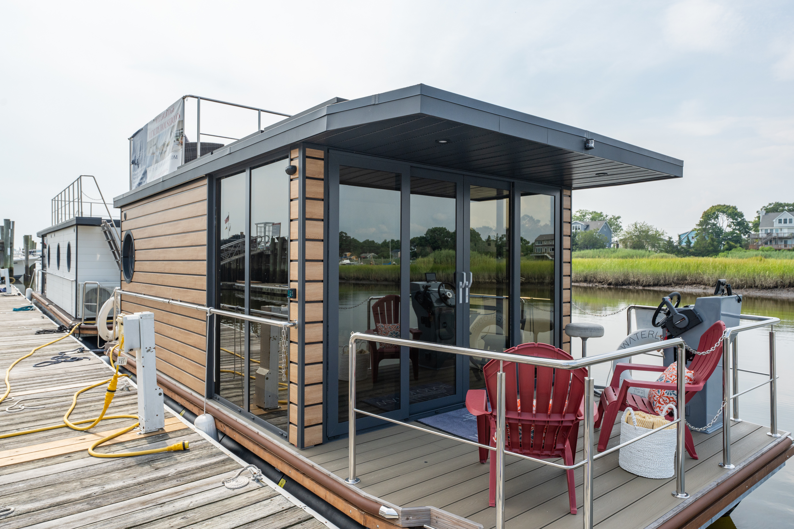 Modern floating house with large windows docked on a calm river, surrounded by nature.